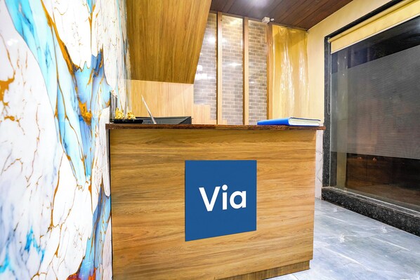 A wooden reception desk with a 'Via' sign, a window with blinds, and a marble wall. - Fabexpress Bala Residency (Mumbai)