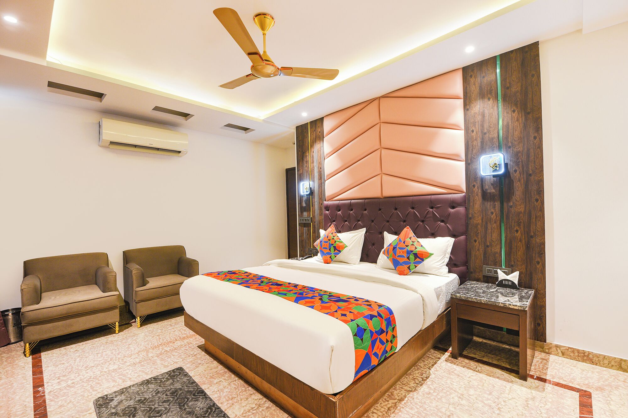 A hotel room with a bed, two armchairs, a nightstand, a ceiling fan, and an air conditioning unit.
