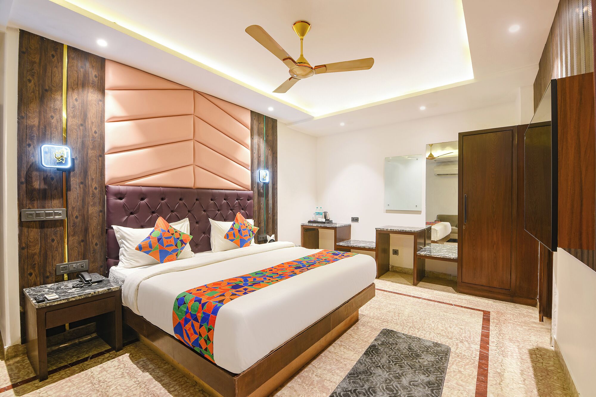 A hotel room with a large bed, a ceiling fan, a bedside table with a phone, a nightstand, a bathroom with a sink and mirror, and a wardrobe.