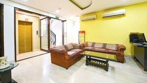 A reception area with a sofa, a coffee table, a desk with a computer, an air conditioning unit, and a staircase leading to an elevator. - Fabhotel Green Park (New Delhi)