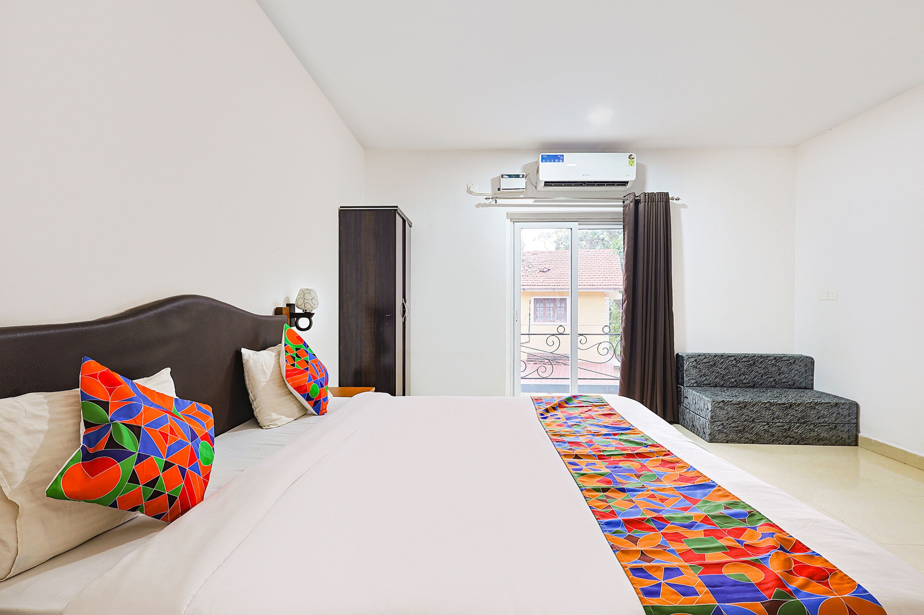 A hotel room with a bed, colorful pillows, a window with curtains, and a balcony with a chair.