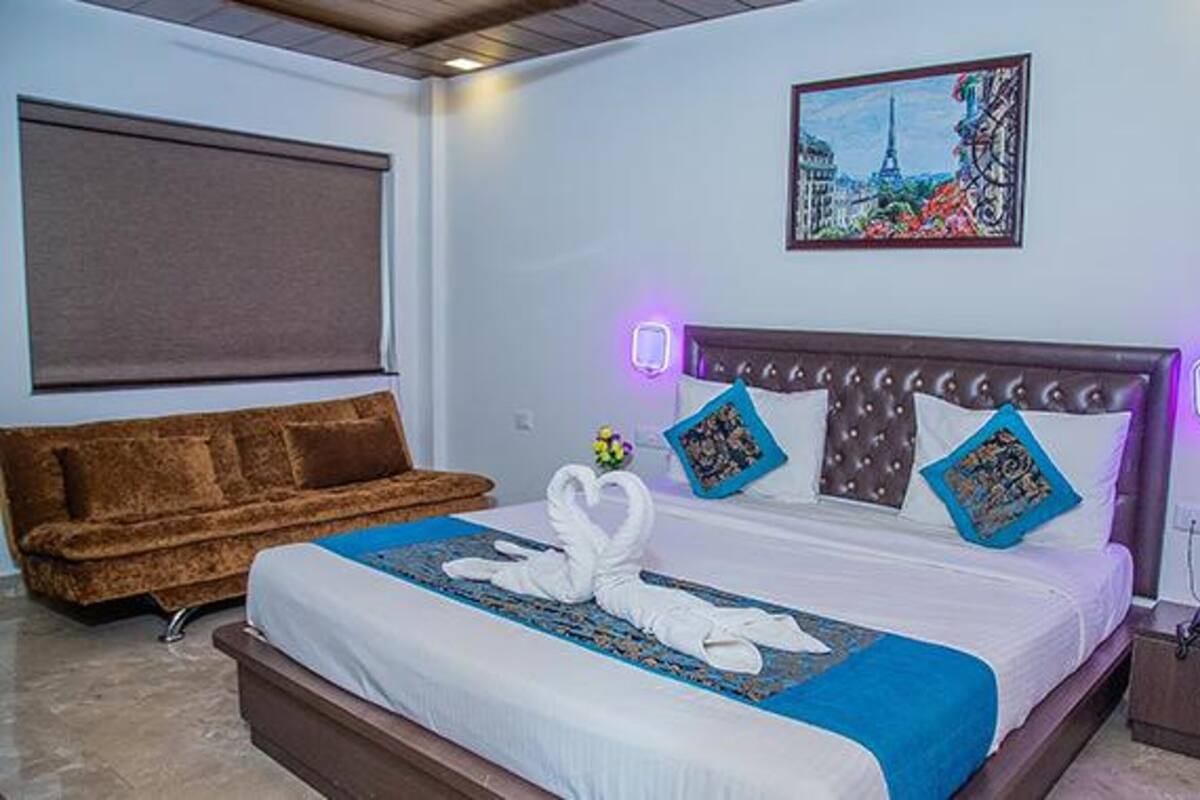 Ein Hotelzimmer mit einem Bett, einem braunen Sofa, einem Stadtbildgemälde und violetten Wandleuchten.