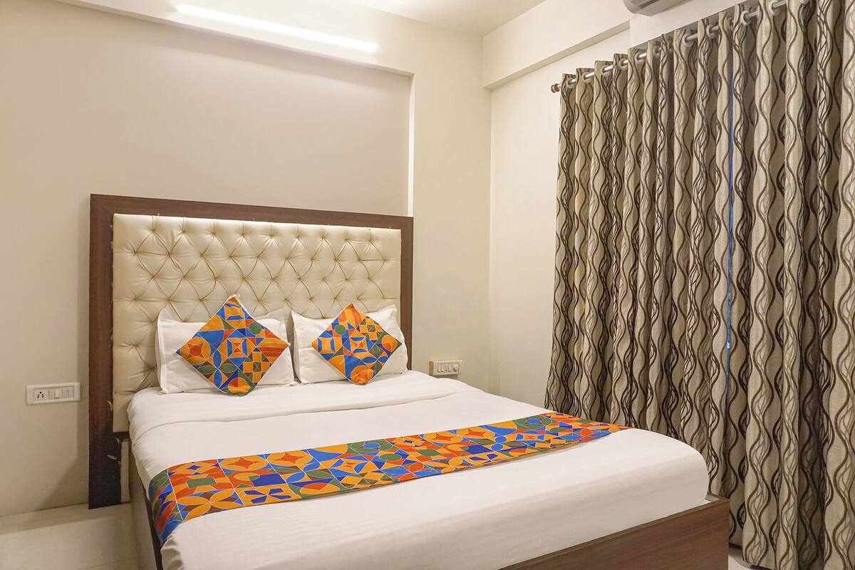 Une chambre d’hôtel avec une tête de lit capitonnée, deux oreillers à motifs géométriques, un lit recouvert d’une courtepointe aux couleurs vives et à motifs géométriques, et un rideau à motifs.