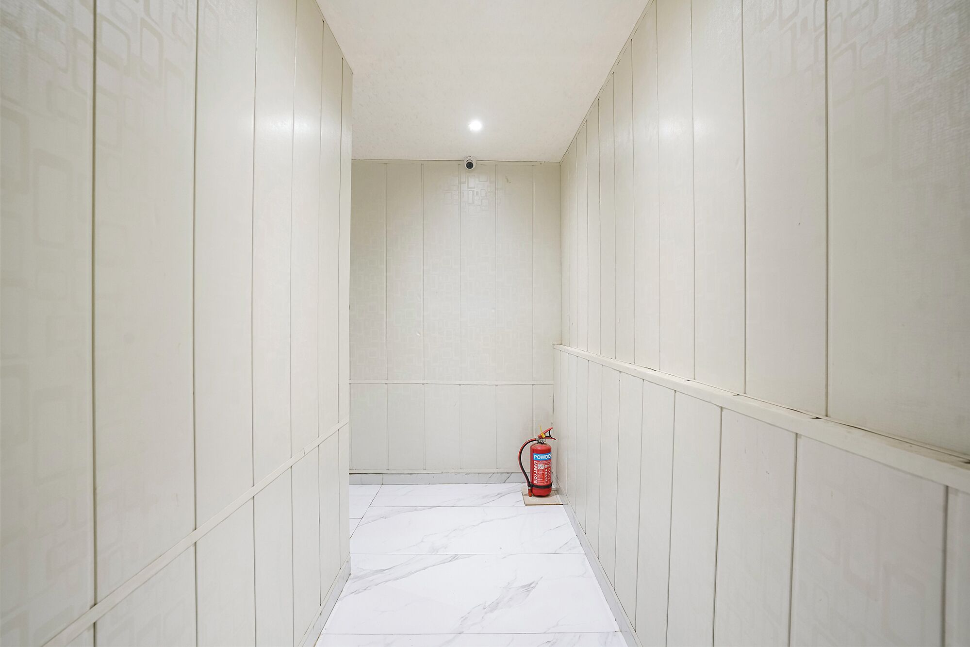 A narrow hallway with white tiled walls and floor, leading to a door at the end.