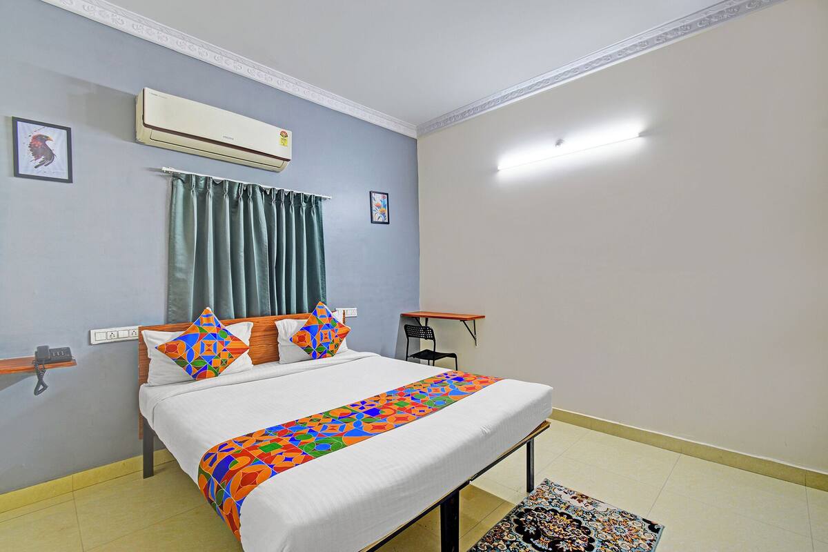 Een hotelkamer met een bed, een bureau en een stoel. Er is een airconditioningapparaat aan de muur en een raam met gordijnen.