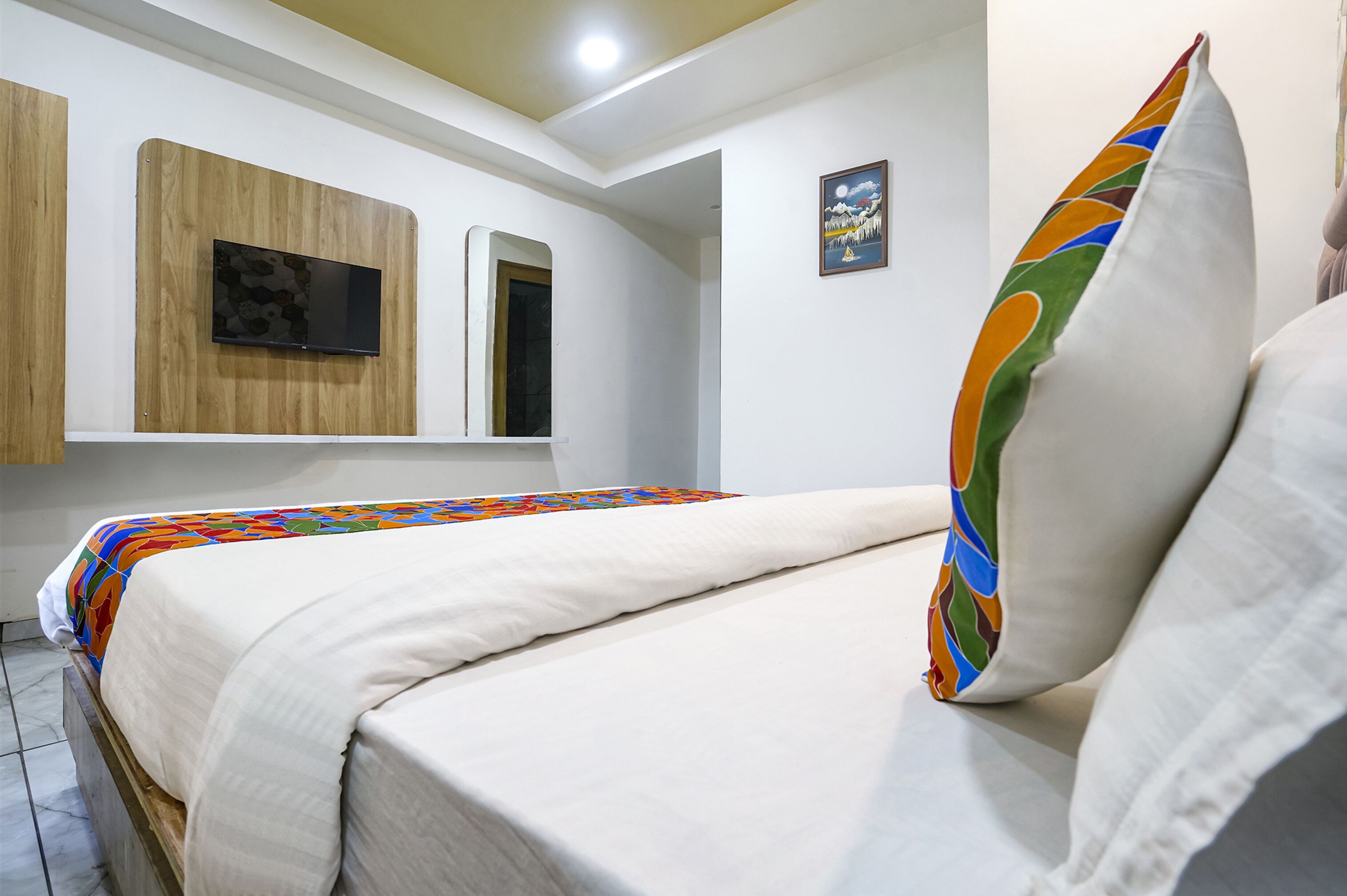 Una cama bien hecha con un edredón colorido, una almohada blanca y otra almohada de colores a la derecha. Hay un televisor de pantalla plana montado en un panel de madera sobre la cama.