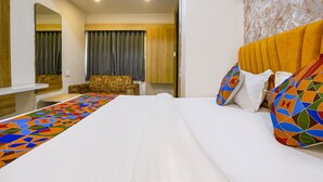 A hotel room with a large bed, a wooden headboard, a colorful throw blanket, and a seating area with a chair and a small table. - FabHotel Alpine I - Inside Shivani Avenue (Ahmedabad)