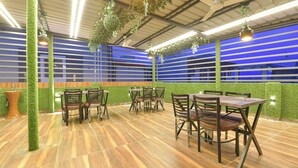 A restaurant with wooden tables and chairs, green artificial plants, and a blue-striped wall in the background. - Fabhotel Mahadev Palace (Haridwar)