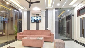 A modern lobby with a pink sofa, a glass coffee table, and a small ottoman. A flat-screen TV is mounted on the wall, and there is a ceiling fan. - Fabhotel Mahadev Palace (Haridwar)