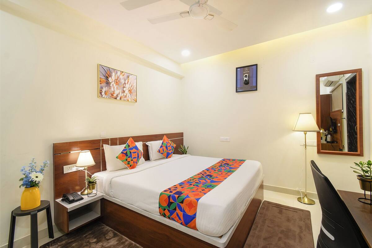 Habitación de hotel con cama, mesita de noche, escritorio, silla, lámpara de escritorio, espejo, ventilador de techo y una pared con dos cuadros enmarcados.