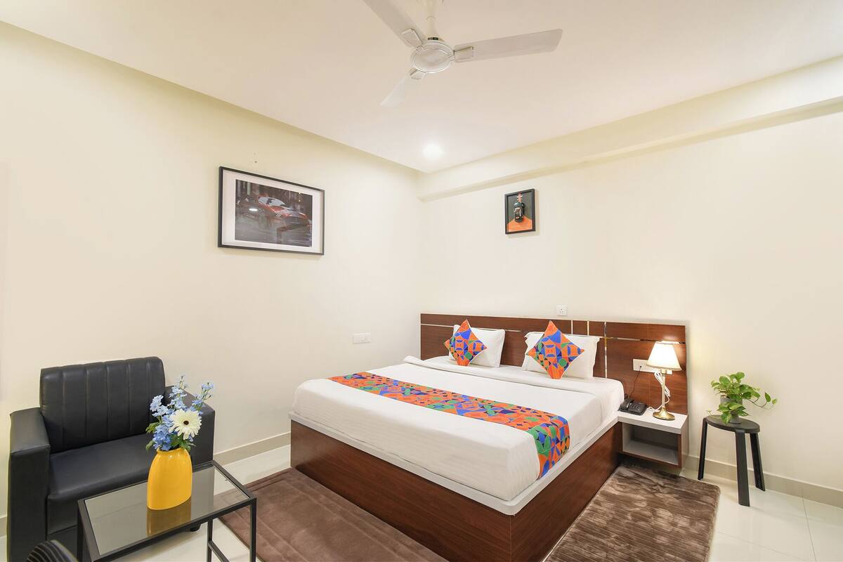 Una habitación de hotel con cama, una silla, una mesita con un florero, una mesita de noche con lámpara y un ventilador de techo.