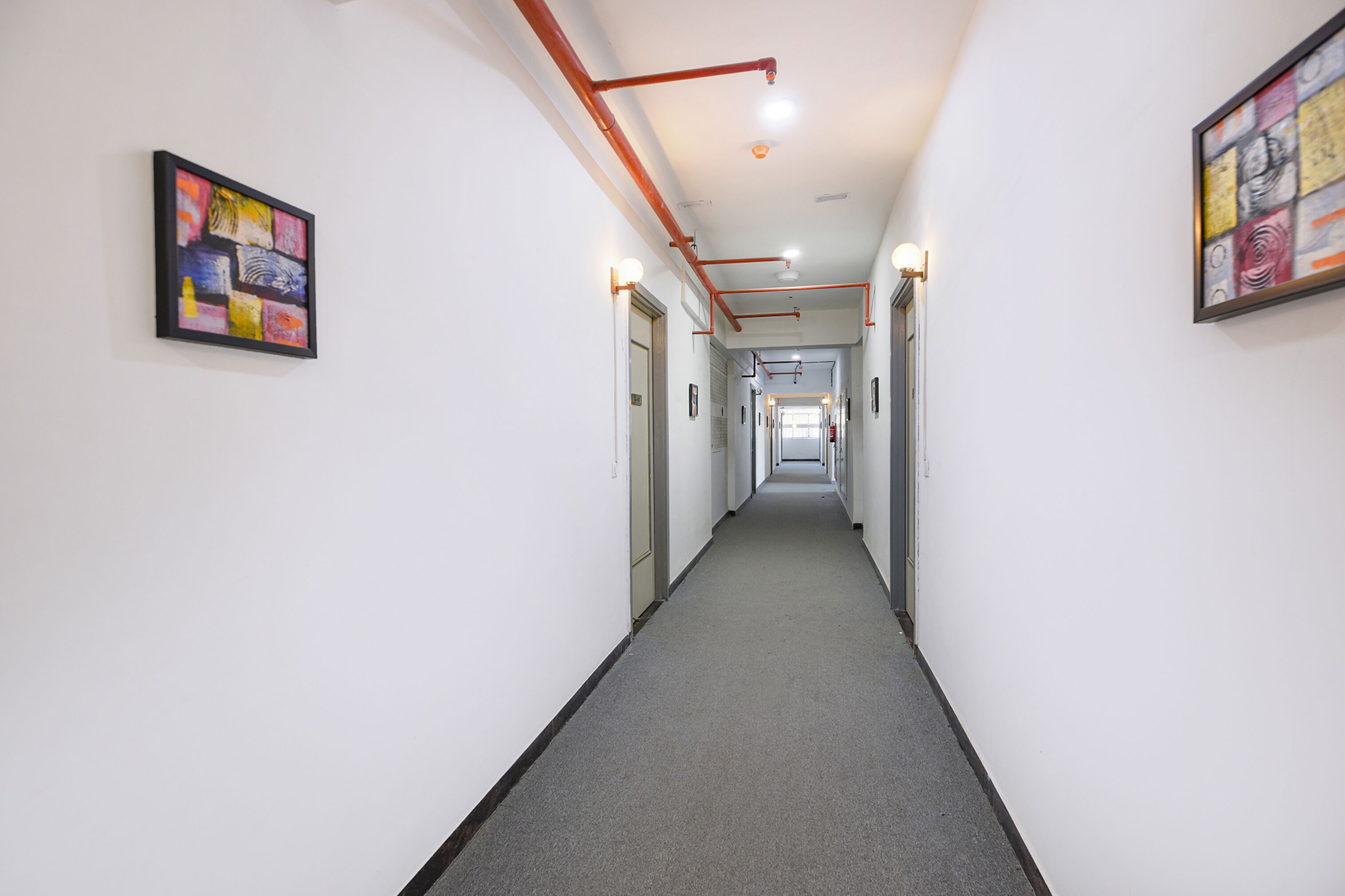 A long, narrow hallway with white walls and a grey carpet. There are two framed artworks on the walls, and the hallway is lined with doors on both sides.