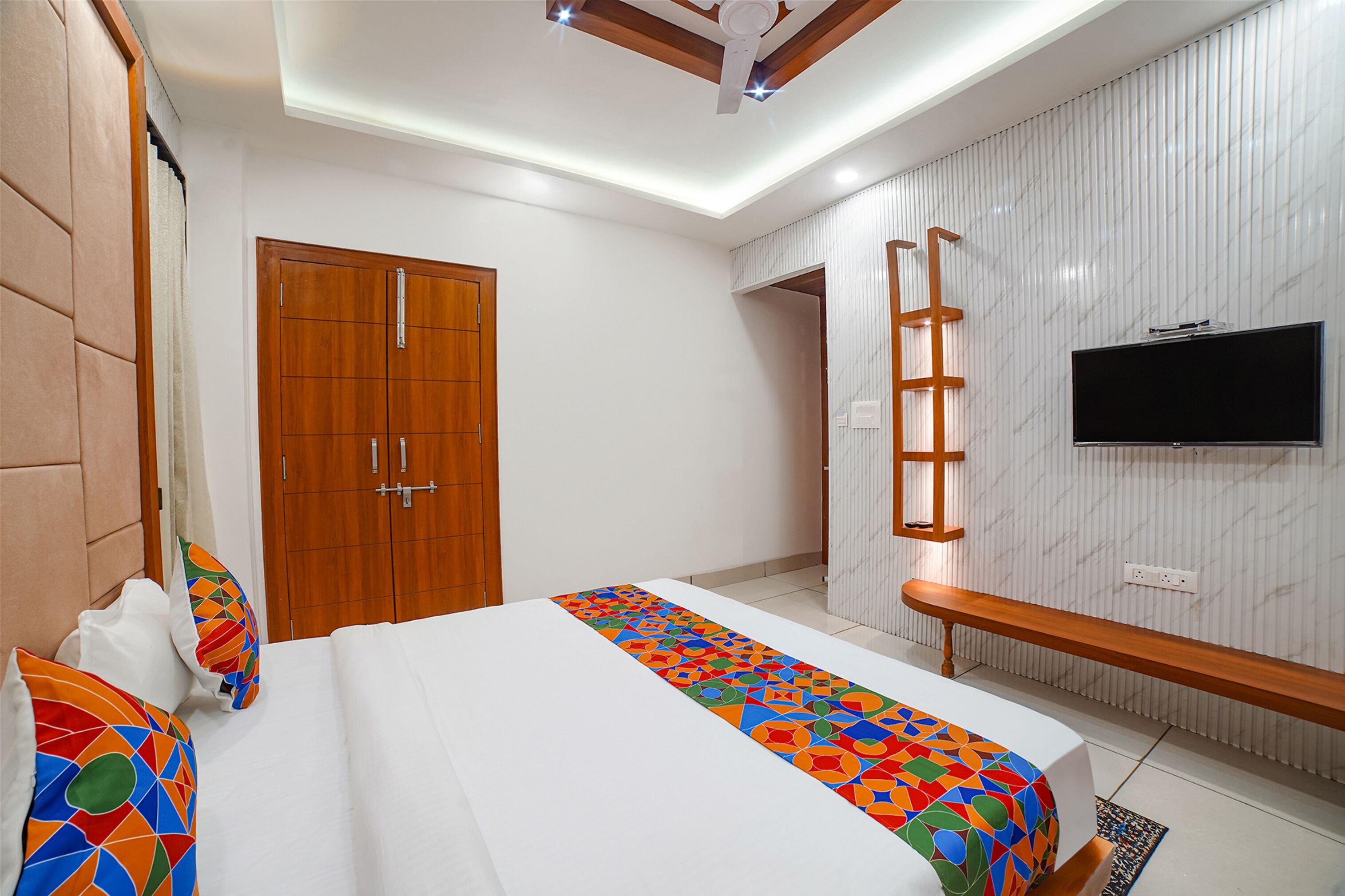 A bedroom with a bed, a wooden wardrobe, a TV mounted on the wall, and a ceiling fan.
