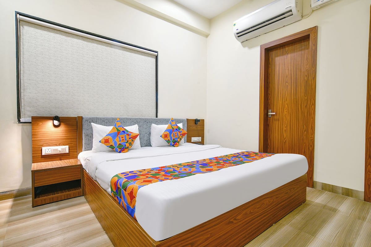 Una habitación de hotel ordenada con un cabecero de madera, una colcha blanca y dos almohadas con motivos geométricos. Hay una mesita de noche de madera con una lámpara, un enchufe y una puerta de madera con manija.