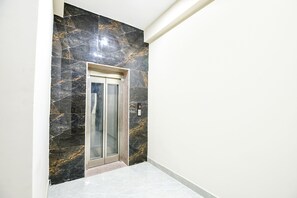 A hallway with a marble wall and a glass elevator door. - Fabexpress Mapple Suites (Faridabad)