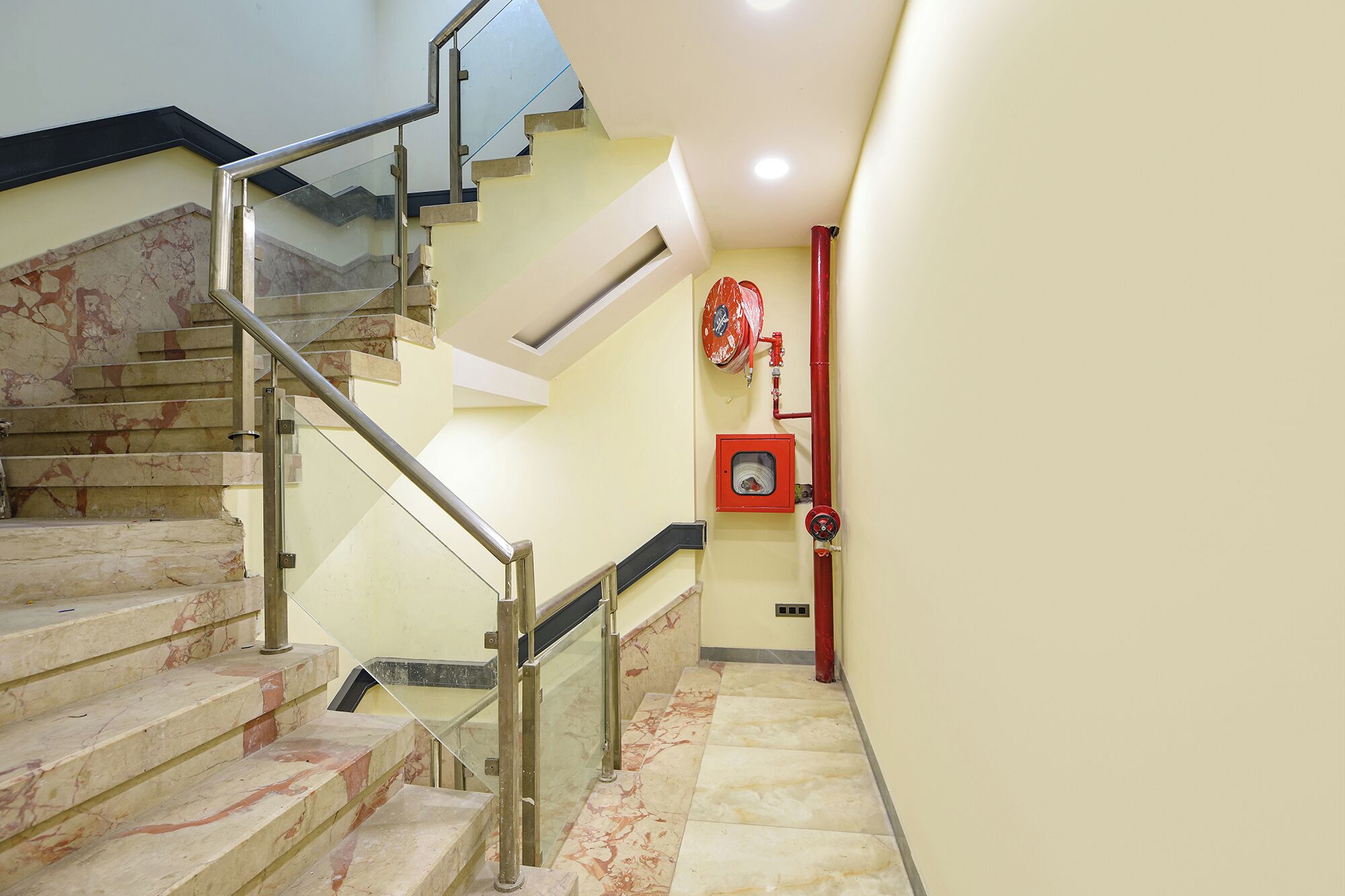Una escalera con escalones de mármol y barandillas de metal, que conduce a una plataforma con una manguera contra incendios roja y un extintor.