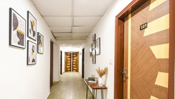 A hallway with framed artwork on the walls, a wooden door with the number 101, and a small table with books and a vase. - Via Krishna Residency - Nr. Nehru Place Metro (New Delhi)
