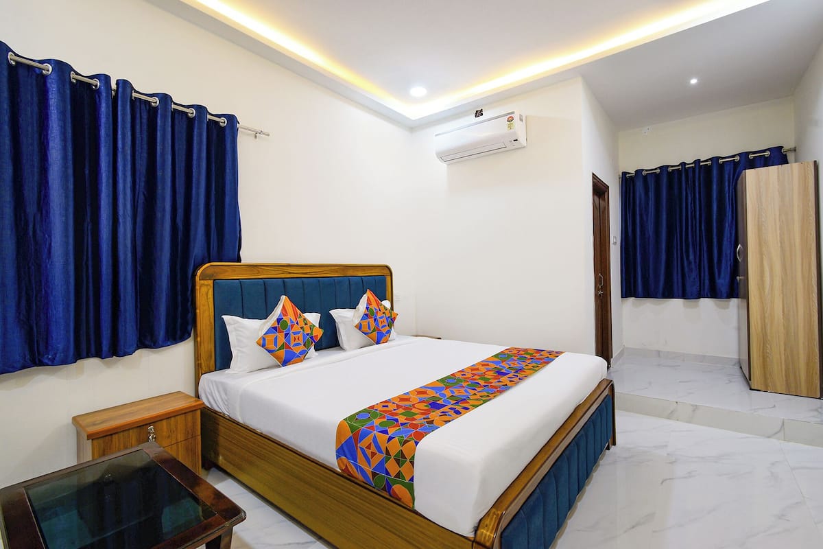 A hotel room with a wooden bed, white bedding, a colorful geometric throw blanket, a nightstand, a wooden wardrobe, and an air conditioning unit on the wall.