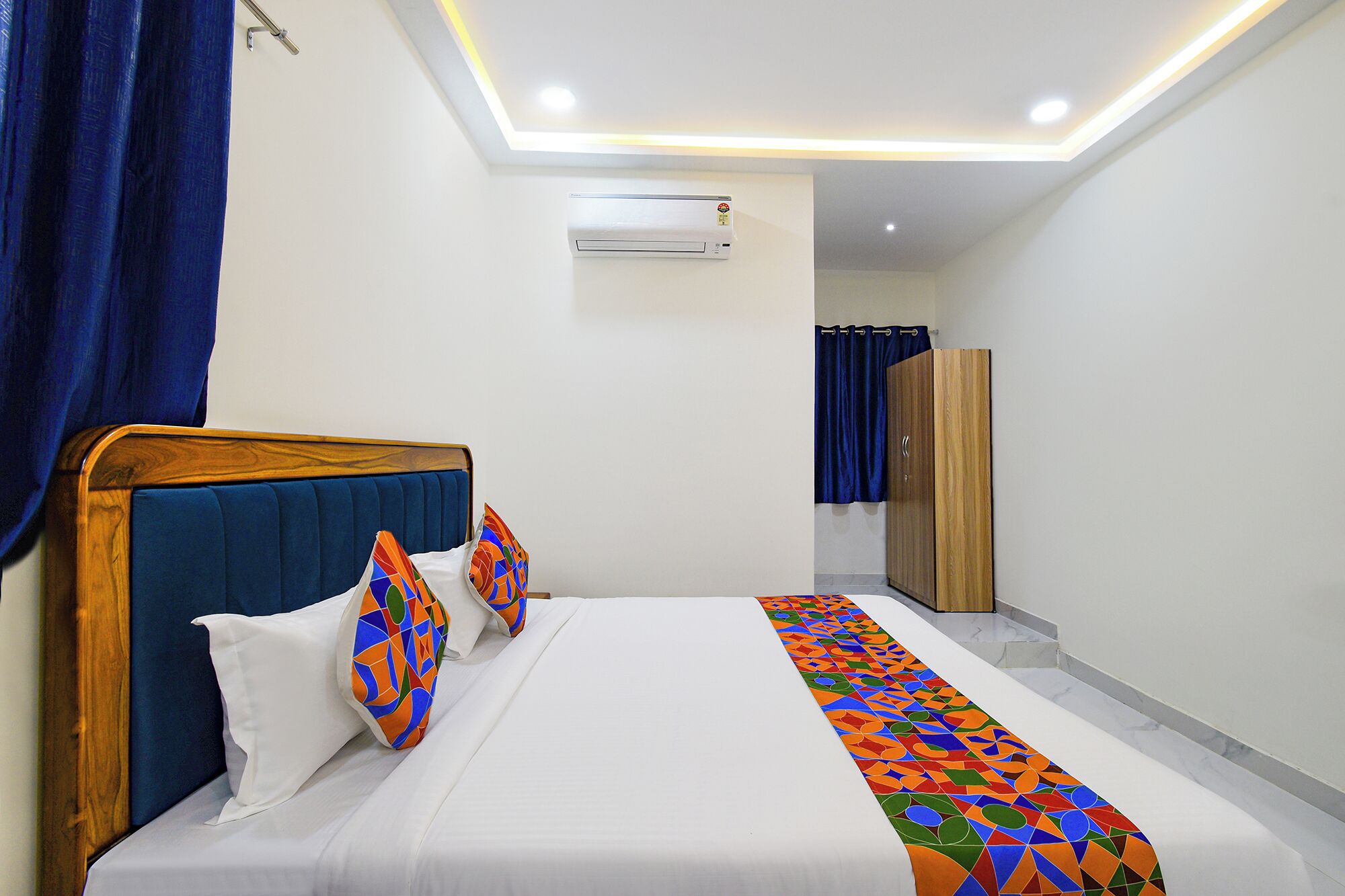 A hotel room with a bed, a blue curtain, a wooden wardrobe, and an air conditioning unit.