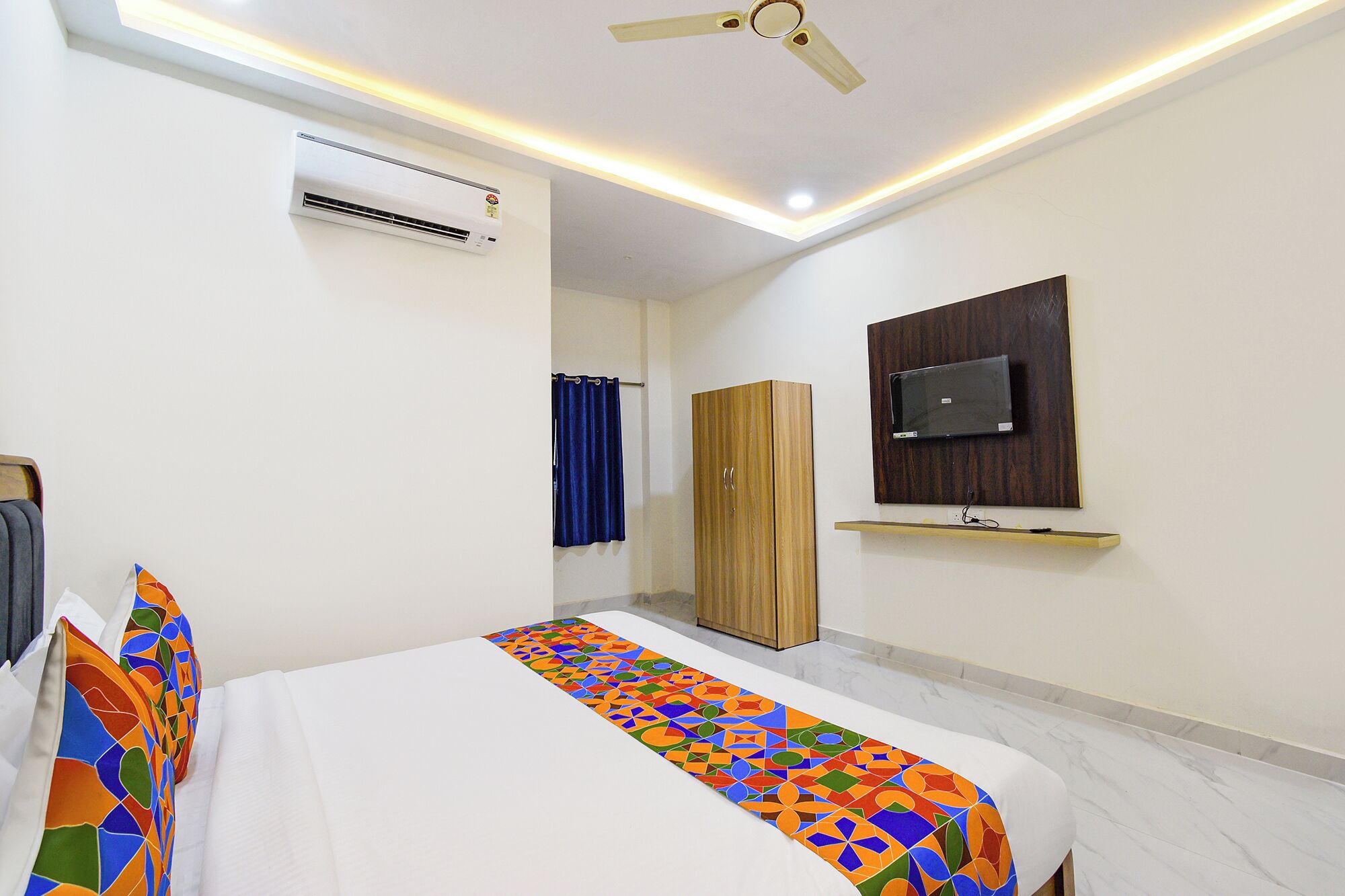 A bedroom with a bed, an air conditioner, a ceiling fan, a wardrobe, a television, and a patterned rug.