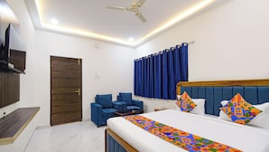 A modern hotel room with a large bed, a seating area with blue armchairs, a ceiling fan, and a TV mounted on the wall.
