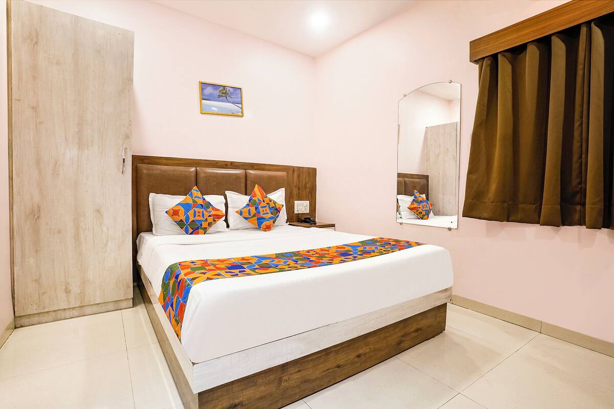 A bedroom with a wooden headboard, a large bed with patterned pillows, a bedside table, a nightstand, a mirror, and a window with curtains.