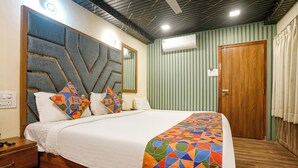 A neatly made bed with a geometric-patterned bedspread, a wooden nightstand with a drawer, a wall-mounted air conditioning unit, and a door with a wooden frame. - FabHotel Malali Residency (Mumbai)