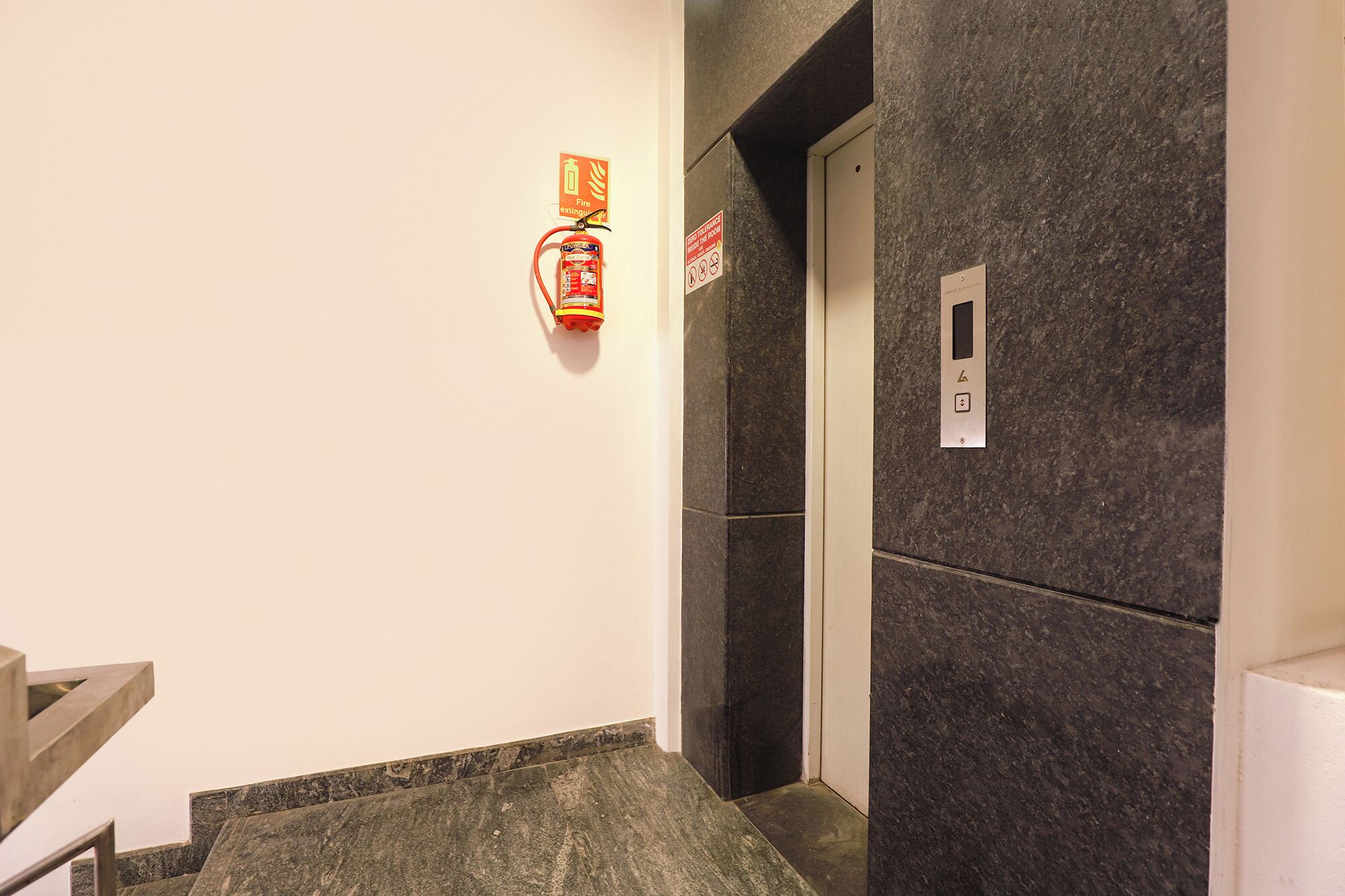 Un couloir avec un extincteur fixé au mur, une porte d’ascenseur et un panneau de commande mural.