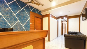 A hotel reception area with a wooden reception desk, a blue patterned wall, a black leather sofa, and a corridor leading to other rooms. - Fabhotel Lloyd’S Inn (Chennai)