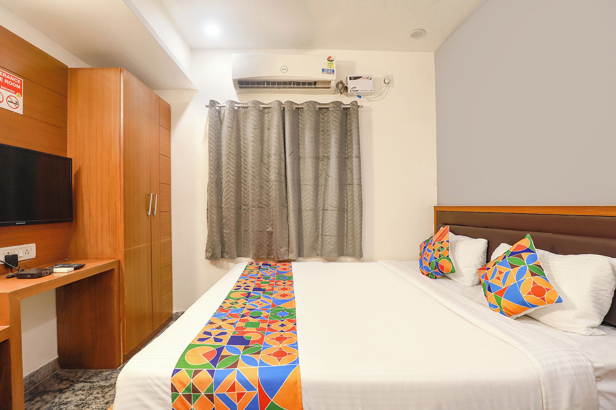 A hotel room with a bed, a TV, a desk, and an air conditioning unit.