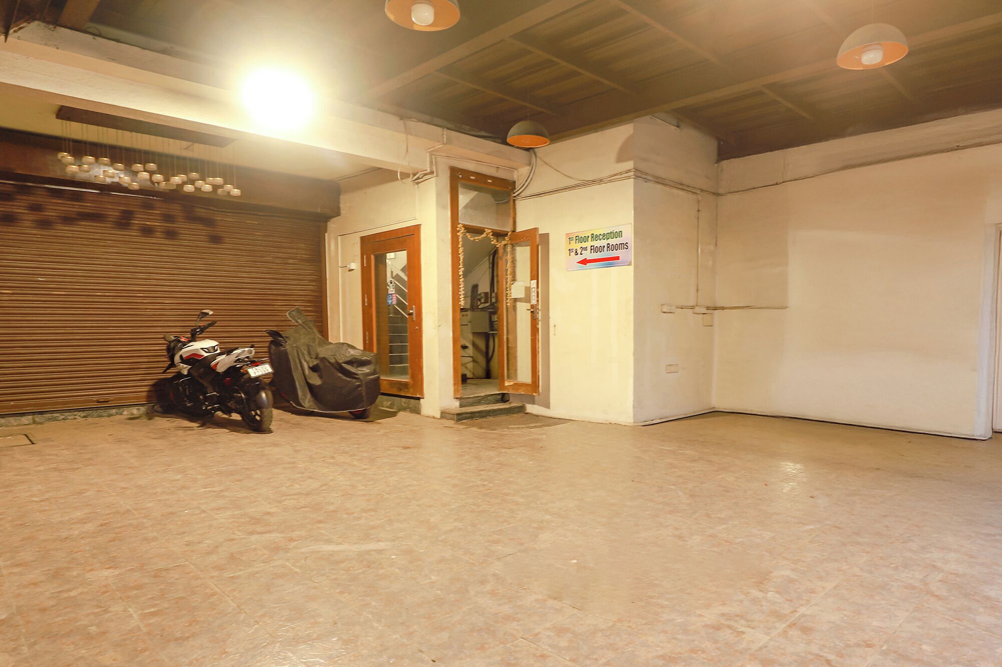 A spacious indoor area with a closed shutter door, a motorcycle, and a sign indicating "1st Floor Reception 1st & 2nd Floor Rooms".