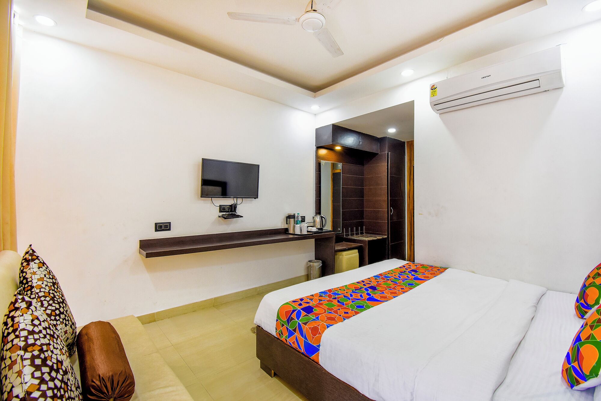 A hotel room with a bed, a TV, a mirror, a shelf with toiletries, and an air conditioner.