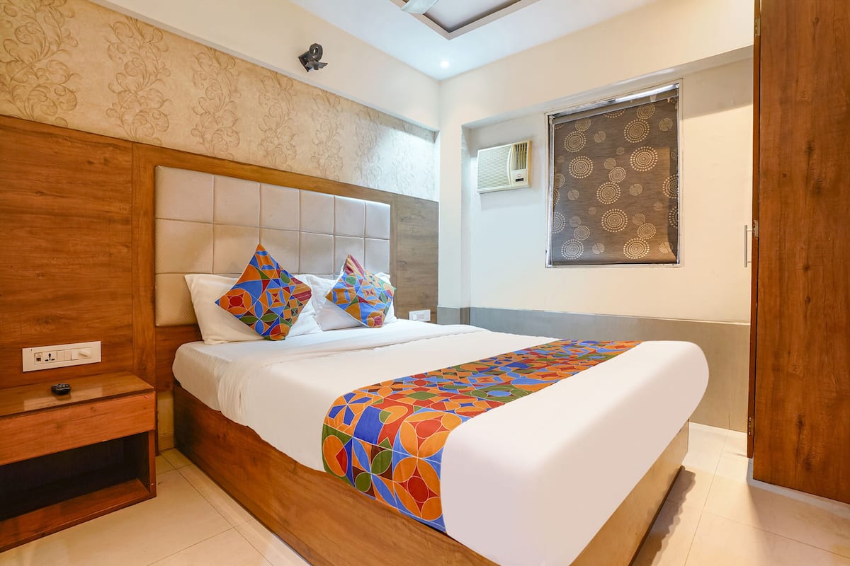 Une chambre d’hôtel avec une tête de lit en bois, un lit avec une literie aux couleurs vives, une table de chevet, un climatiseur fixé au mur et une fenêtre avec un store à motifs.