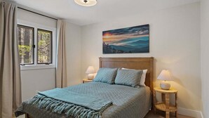 A bedroom with a bed, two bedside tables, a window with curtains, a painting on the wall, and a ceiling light.
