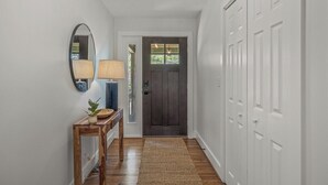 A hallway with a wooden floor, a dark door, white walls, a mirror, a lamp, and a small table with a plant.