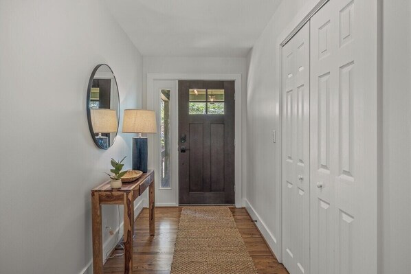 A hallway with a wooden floor, a dark door, white walls, a mirror, a lamp, and a small table with a plant.