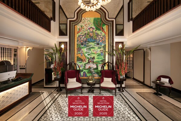 A hotel lobby with a large mural, two red upholstered chairs, a reception desk, and a floor with a geometric tile pattern. - La Mejor Indochine Hotel (Hanoi)