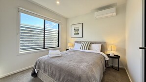 Un dormitorio con un ventanal grande, una cama con manta gris, dos mesitas de noche con lámparas y un aire acondicionado de pared.