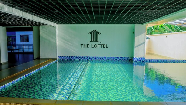 Une piscine intérieure avec un sol et des murs carrelés, un plafond à motifs géométriques et le logo de « The Loftel » sur le mur.