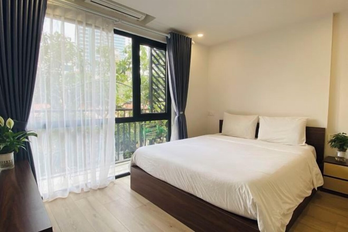 A bedroom with a large bed, a wooden floor, a window with curtains, and a small bedside table with a plant.