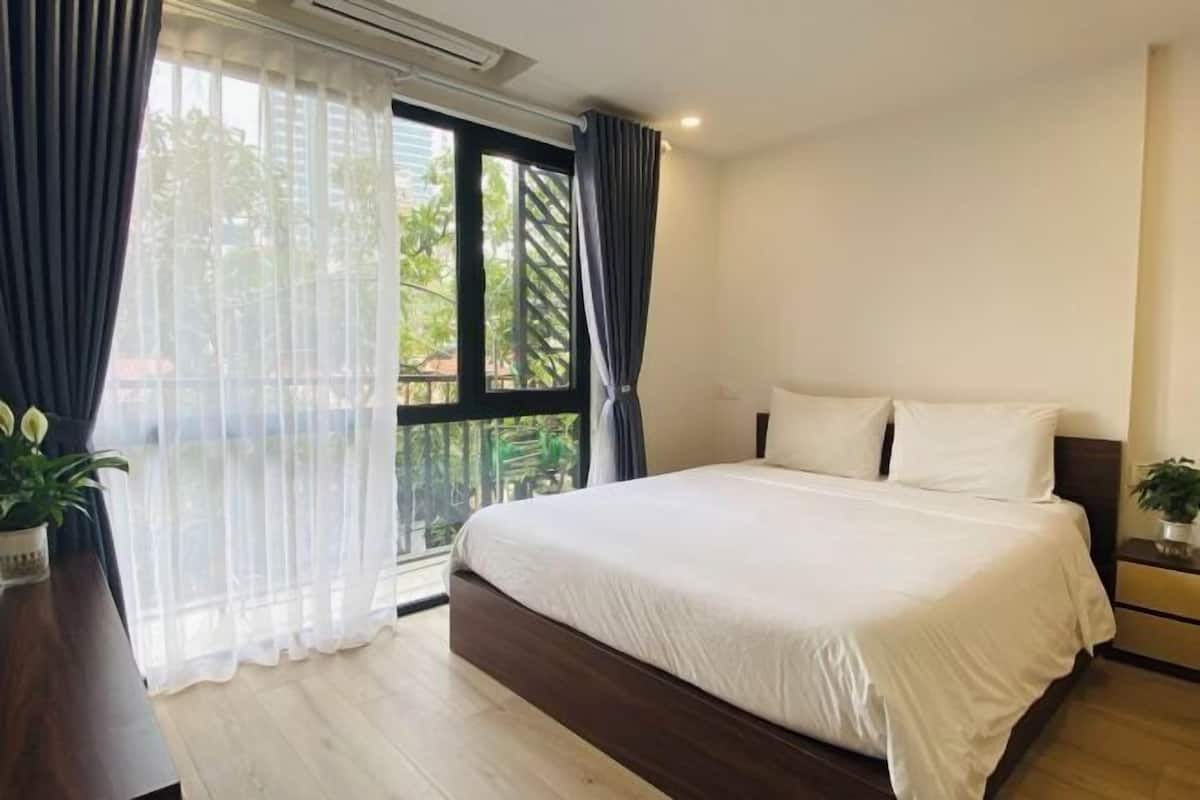 A bedroom with a large bed, a wooden floor, a window with curtains, and a small bedside table with a plant.