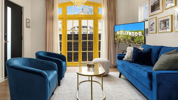 Una sala de estar con un sofá azul, dos sillones azules, una mesa de centro de vidrio y una araña. Un televisor grande en la pared muestra el cartel de Hollywood.