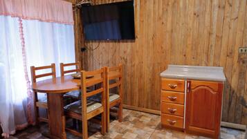 Une pièce aux murs lambrissés, avec une table et des chaises en bois, un petit coin cuisine doté d’un plan de travail et de tiroirs, et un téléviseur à écran plat fixé au mur.