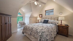 Een slaapkamer met een schuine plafond, een bed met bloemenbedekking, een nachtkastje met lampen en een raam met uitzicht op groen.