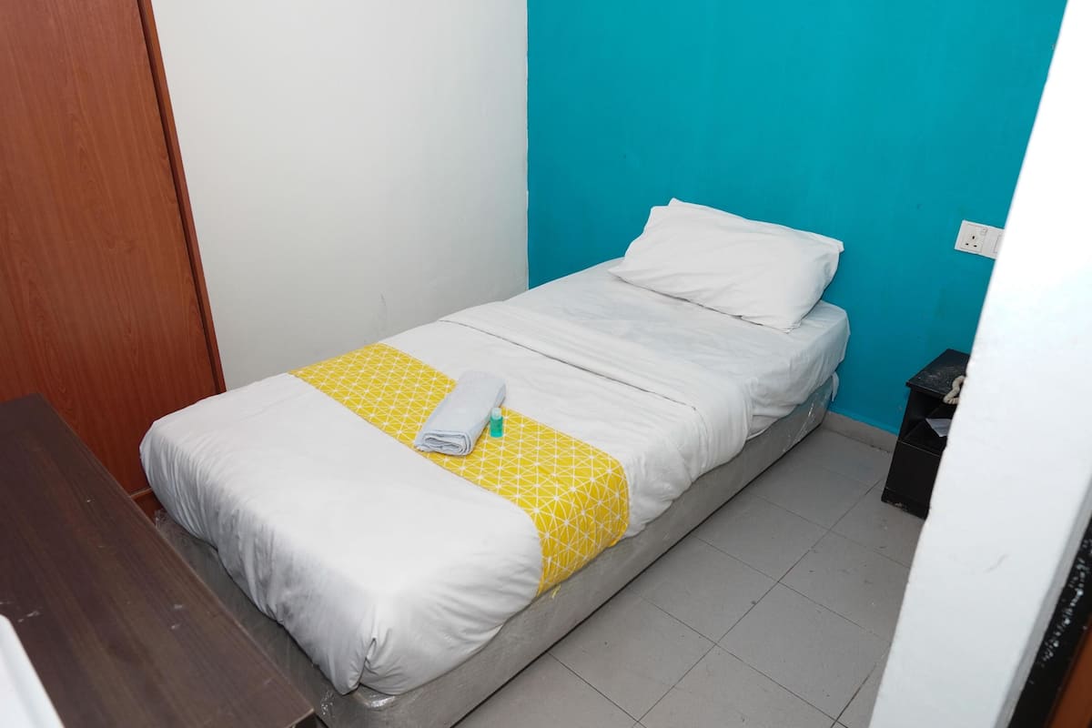 Een enkel bed met een gele, gemêleerde spreideken en witte kussens, een houten nachtkastje en een zwarte prullenbak in een kleine kamer met wit- en blauwe wanden.