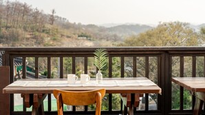 Una mesa de madera con dos tazas blancas y una botella de vidrio transparente en un balcón con vistas a las colinas.