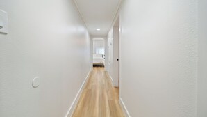 Un couloir étroit aux murs blancs et au plancher en bois, qui mène à une pièce comprenant un lit et une fenêtre avec des rideaux.