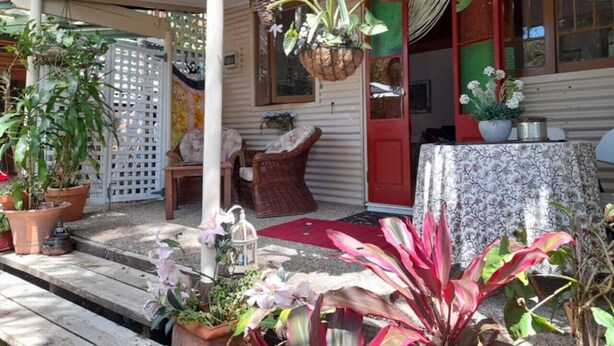 A covered porch with potted plants, wicker furniture, and a red door. - Quirky Cottage in Centre of Maleny Walk Everywhere (Maleny)