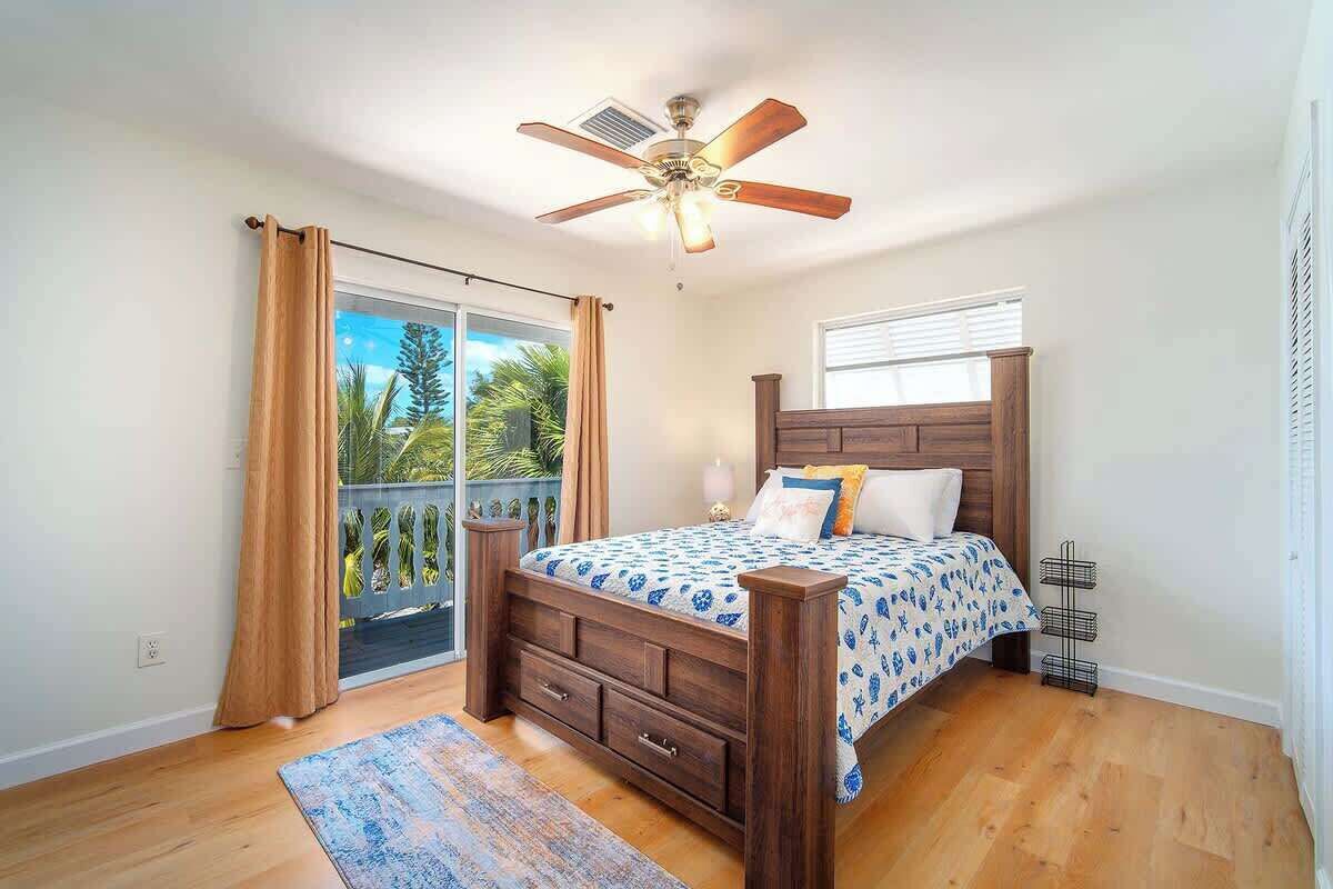 A bedroom with a wooden bed, a ceiling fan, a large window with curtains, and a view of greenery outside.