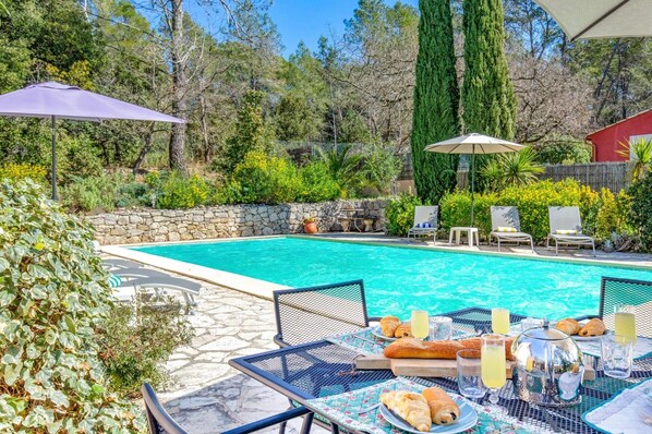 A swimming pool with lounge chairs and umbrellas, surrounded by greenery and a stone wall.